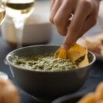 Dłoń macza nachosa w miseczce guacamole, w tle kieliszki wina
