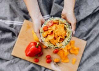 Domowe nachos z piekarnika z serem, fasolą i guacamole: imprezowa przekąska jak z food trucka Dłonie trzymające nachos z guacamole i pomidorkami na desce